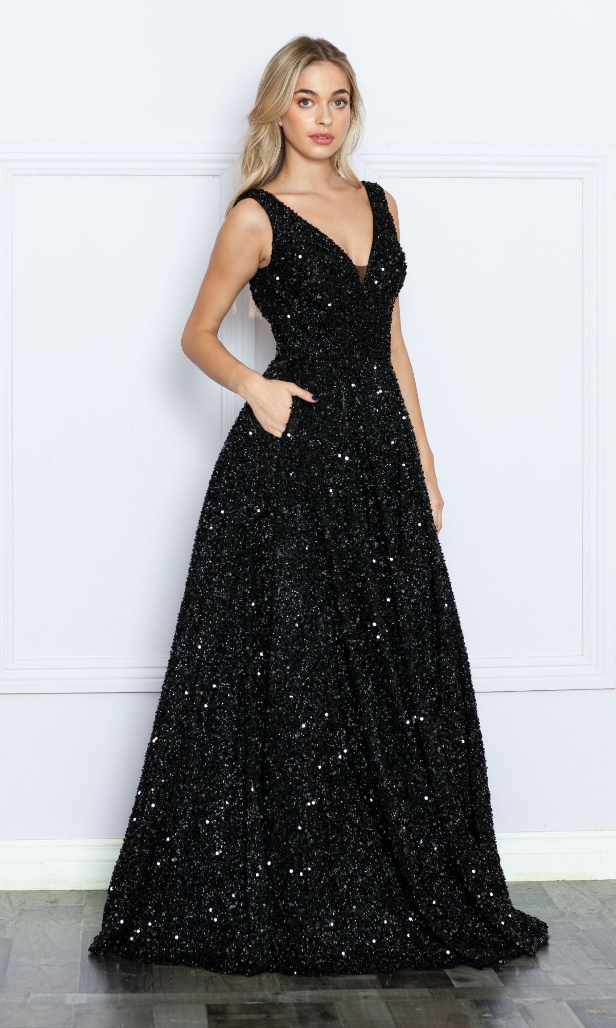 A woman stands indoors against a plain white wall, wearing the Long Black Sequin A-Line Prom Dress 9168—a sleeveless gown with a deep V-neckline, pockets, and a floor-length A-line silhouette.